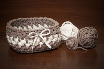 Fototapeta premium Knitted basket of handmade knitted yarn on a wooden background. Around the basket are balls of knitted yarn in white and brown.