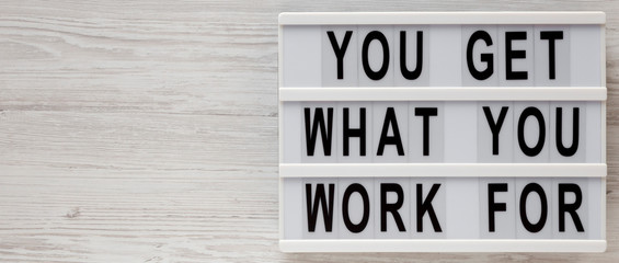 'You get what you work for' words on a lightbox on a white wooden surface, top view. Overhead, from above, flat lay. Copy space.