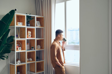 Young Asian man having a cup of coffee while looking out of the window at home.