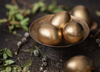 Golden easter eggs are scattered on a dark background with dry green branches. eggs in a golden bowl.