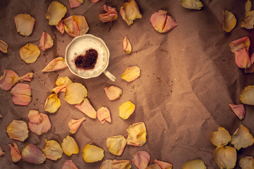 Coffee mug with cream and cinnamon in the form of a heart with pink flower petals on crumpled kraft paper. Flat lay. Copy space. Vignetting.