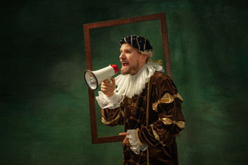 Calling for sales. Portrait of medieval young man in vintage clothing with wooden frame on dark background. Male model as a duke, prince, royal person. Concept of comparison of eras, modern, fashion.