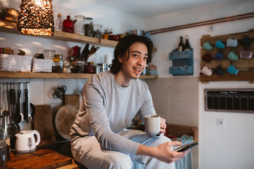 young man using mobile phone and drinking coffee in his kitchen