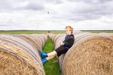 Naklejka na ściany i meble Child playing on long row of round hay bales on overcast day. Childhood on the farm, country life.