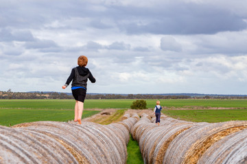 Children running on long row of round hay bales on overcast day. Childhood on the farm, country life.