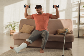 Fototapeta na wymiar Smiling man with broken leg exercising with dumbbells on couch in living room