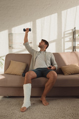 Fototapeta na wymiar Handsome man with leg in plaster bandage training with dumbbell and holding remote controller on couch