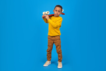Fototapeta na wymiar Funny little African-American boy with a skateboard smiles and looks away, standing on a blue background. Concept of activity and happy childhood.