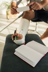 Cropped view of man taking photo with smartphone while holding pencils in broken leg near notebook on ottoman