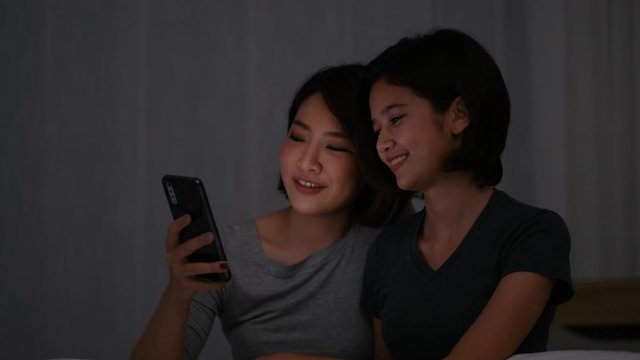 Couple Asian teen girls look to mobile phone and talk together in bedroom at night.