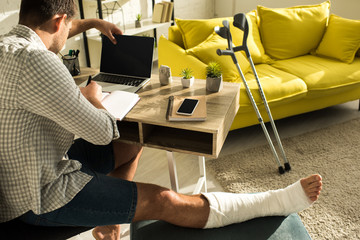 Back view of freelancer with broken leg working with laptop and notebook at table