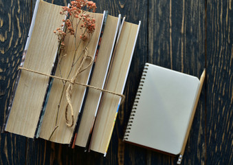 A stack of books tied with twine, dried flowers and notebook with pencil on a dark wooden background. Top view. Close-up. Flat lay.