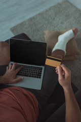 High angle view of man with leg in plaster bandage using laptop and holding credit card