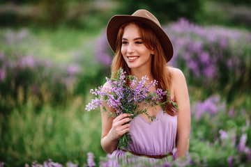 Red-haired girl in a hat lies in the grass with purple flowers. Young woman smile in nature. lady...
