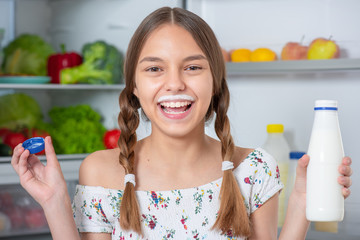 Beautiful young teen girl holding bottle of milk and drinks while standing near open fridge in kitchen at home. Portrait of pretty child choosing food in refrigerator full of healthy products. - 322308466
