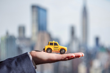 The hand of a businessman in a suit holding a yellow toy taxi car on the background of New York skyscrapers. Concept. Taxi city service