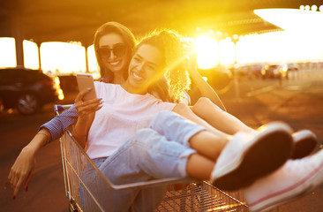 Two young girls having fun on shopping trolleys in summer day with sunlight. Young friends having fun and take selfie on the phone together.  Lifestyle, leisure, entertainment, youth concept.