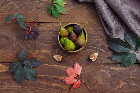 Fruits whole and cut figs in a wooden bowl on a brown wooden background with autumn leaves and blue berries of wild grapes and a brown napkin view from the top