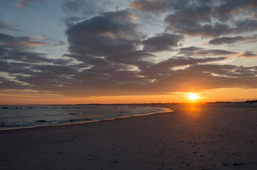 Naklejka na ściany i meble The sun sets behind some clouds along the coastline of a sandy beach with a colorful sky.
