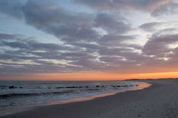 The sun sets behind some clouds along the coastline of a sandy beach with a colorful sky.