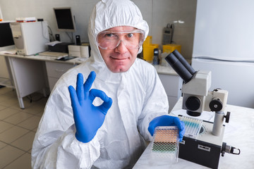 A biologist in a white protective suit and glasses works with a microscope in the laboratory. The...