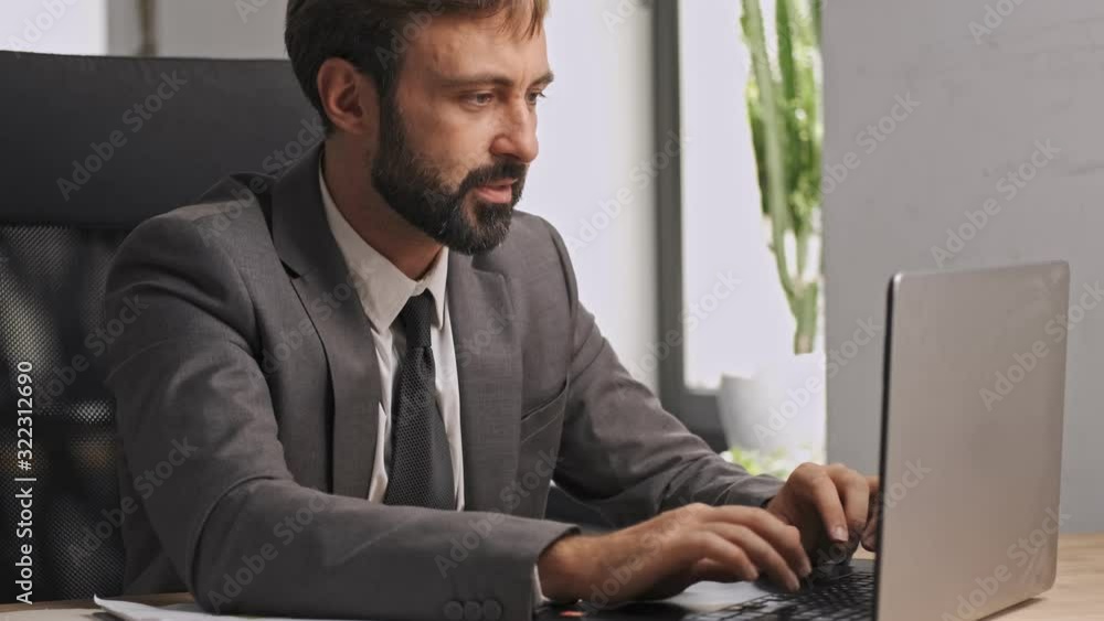 Wall mural Concentrated businessman in formal suit typing something in laptop computer while sitting by the table in office