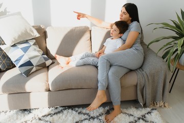 Cheerful mother sitting on couch in cozy light living room holds on lap tickles of little son enjoy...