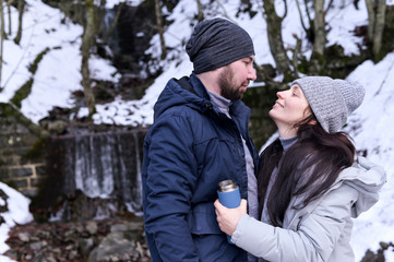 Young woman and man cuddling in a snowy forest. A pair of lovers in winter clothes. The girl holds a thermos in her hands. Copy space.