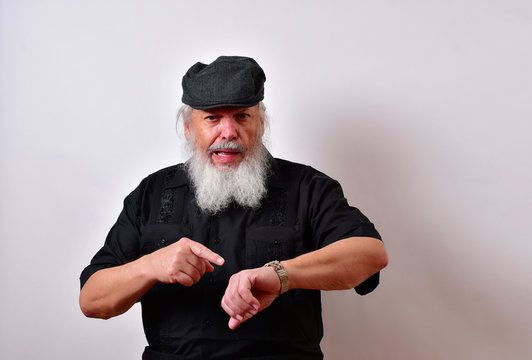 Impatient old man with a scowl on his face pointing at his watch. .. .Mature gentleman with a newsboy cap and black guayabera shirt and long white beard..