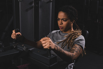 selective focus of tattooed african american girl with dreadlocks working out on fitness machine