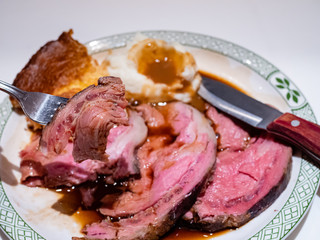 Close up of medium-rare roast beef on a fork. A Sunday roast - slices of roast beef and gravy, mashed potato and Yorkshire pudding - sits in the background