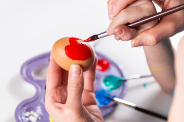 Mother and her child hands painting Easter eggs. Mother helps her son paint an egg with a paintbrush at home. Hand draws a red heart. Happy family preparing for Easter