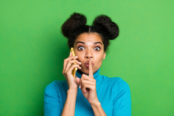 Fototapeta na wymiar Photo of funny dark skin lady hold telephone speaking with colleagues hold finger on lips mouth asking keep silence wear blue turtleneck isolated green color background