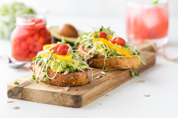 Two pieces of avocado toast with a glass of kombucha in behind and a jar of baby sweet peppers for...