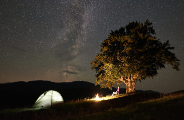 Happy female hiker having a rest at summer night camping in the mountains beside campfire and glowing tourist tent, sitting on chair under big tree and beautiful night sky full of stars and Milky way.