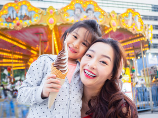 happy asia mother and daughter have fun in amusement carnival park with farris wheel and carousel...
