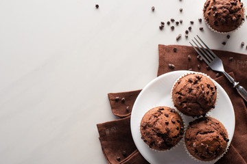 top view of fresh chocolate muffins on white plate and brown napkin near fork on marble surface