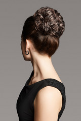 Close-up back shot of a brown-haired lady with a fancy hairdo made as a lustrous hair bun. The girl with a long neck is posing on the gray background.