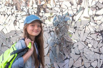 Young girl on the background of a broken mirror and smiling. Girl schoolgirl in a cap and with a backpack