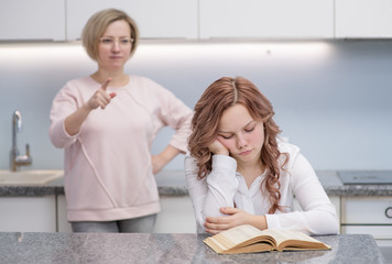 A teenage girl is sitting upset at a table in her kitchen. Her mother stands in the background, she...
