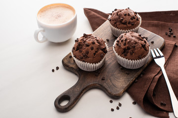 fresh chocolate muffins on wooden cutting board near napkin, fork and coffee