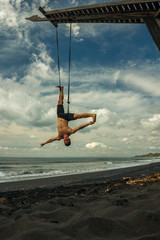 outdoors portrait of young attractive and athletic man practicing aero yoga workoutat the beach hanging from rope swiing above the sea training body balance and flexibility