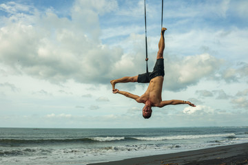 outdoors portrait of young attractive and athletic man practicing aero yoga workoutat the beach hanging from rope swiing above the sea training body balance and flexibility