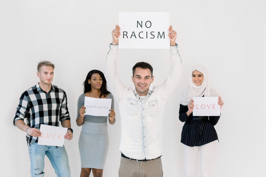 Stop racism, no racial discrimination of people. Young Caucasian man holds a poster with No racism text, while standing together with multiethnical friends with slogans on white background indoors