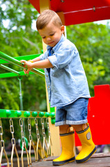 A little cute three-year-old boy plays on the playground and slides down the slides in yellow rubber boots.	