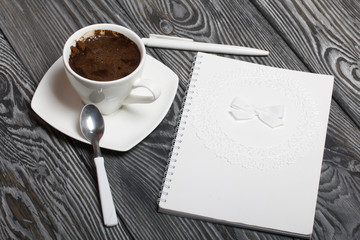 Notebook white with a spiral. The cover is decorated with paper lace and a ribbon tied to a bow. Nearby lies a white pen. It is coffee in a white cup on a saucer. On brushed pine boards.