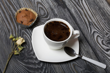 Coffee in a white cup stands on a saucer. Nearby lies a muffin, and a dried flower of a beige rose. On brushed pine boards.