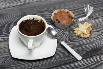 Coffee in a white cup stands on a saucer. Nearby lies a muffin, and a spoon on a saucer. In a bag a set of cookies in the form of snowflakes. On brushed pine boards.