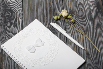 Notebook white with a spiral. The cover is decorated with paper lace and a ribbon tied to a bow. Nearby lies a white pen and a dried flower of a beige rose. On brushed pine boards.