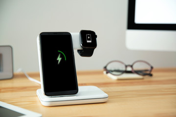 Mobile phone and smartwatch with wireless charger on wooden table. Modern workplace accessory - obrazy, fototapety, plakaty